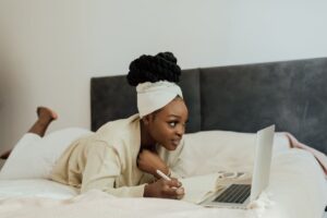Young woman in headwrap working remotely with laptop and notebook from bed, showcasing a relaxed lifestyle.