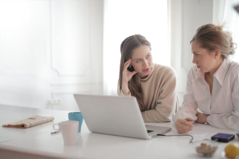 Two women engage in a focused work discussion at a desk, utilizing a laptop in a bright office space.