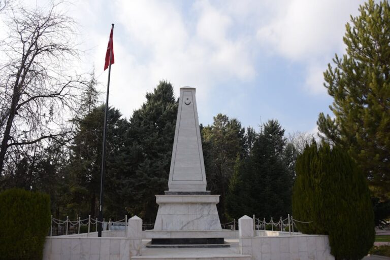 A tall stone obelisk monument in a park with surrounding trees and a national flag.