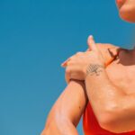Close-up of a woman applying sunscreen to her shoulder with a tattoo visible in a bright, sunny setting.