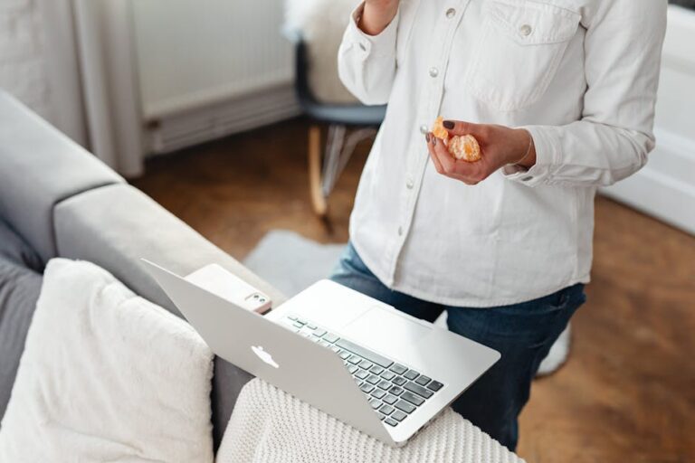 Person working from home, snacking on an orange while using a laptop in a cozy living room.